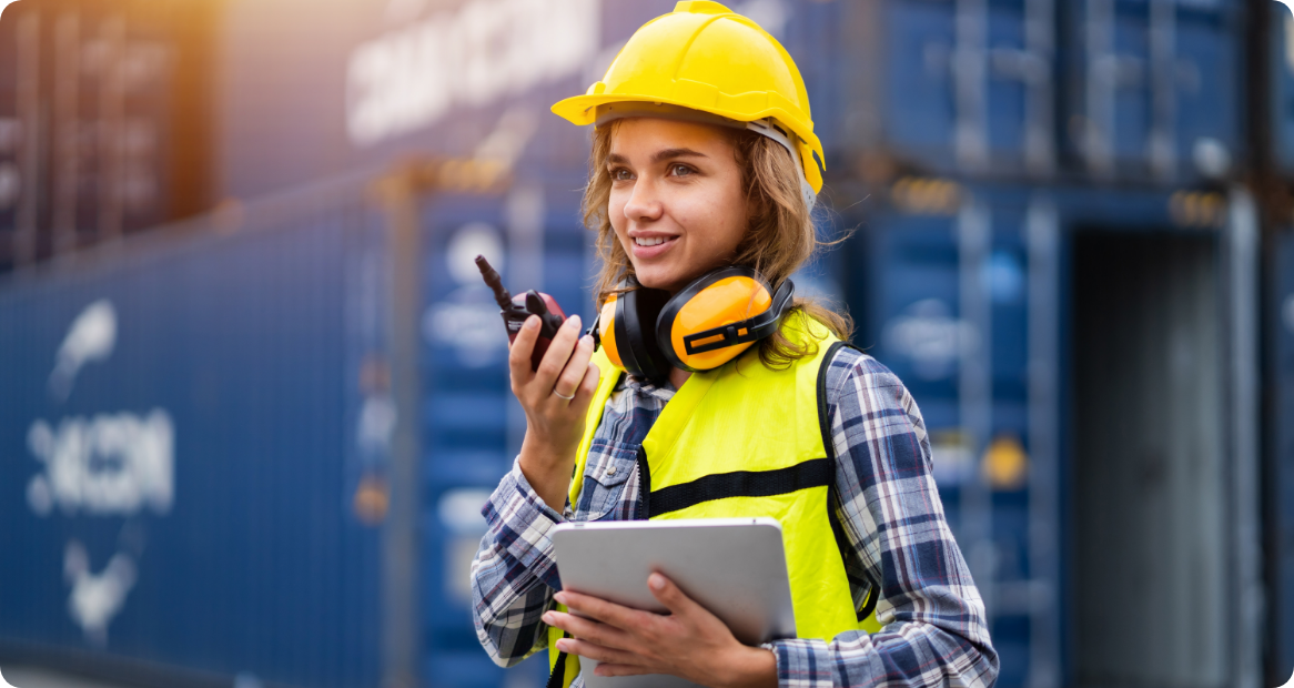 A female logistics worker using a walkie-talkie at a shipping container port.