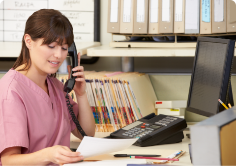 A female medical receptionist in pink scrubs talking on the phone at a clinic desk.